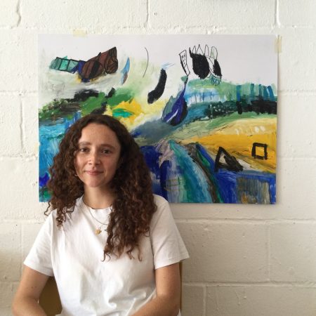 A photo of Gabby Jonbes. A white woman with long dark curly hair. She is wearing a white t shirt and is sitting n front of a painting by Ali Kamalati.