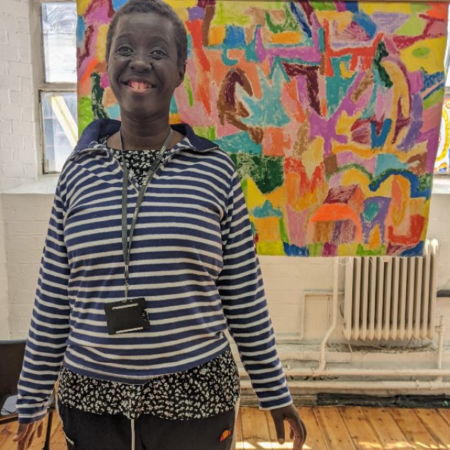 A woman wearing a striped hoodie smiling at the camera, in front of her artwork.