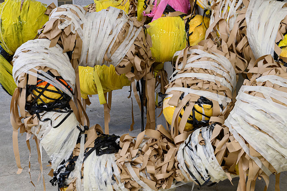Close up of a large abstract sculpture, yellow, beige, white, black, and pink tape wrapped tightly together.