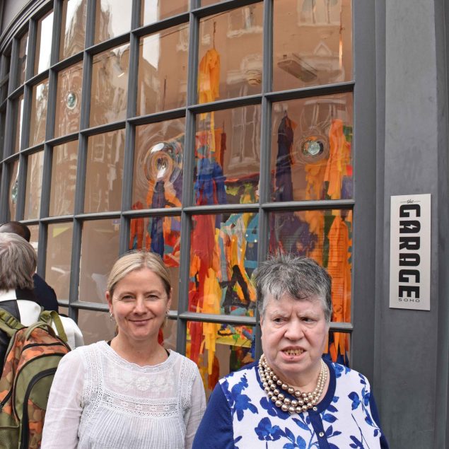 Image of two women posing for a photograph together in front of a window gallery on a street outside.