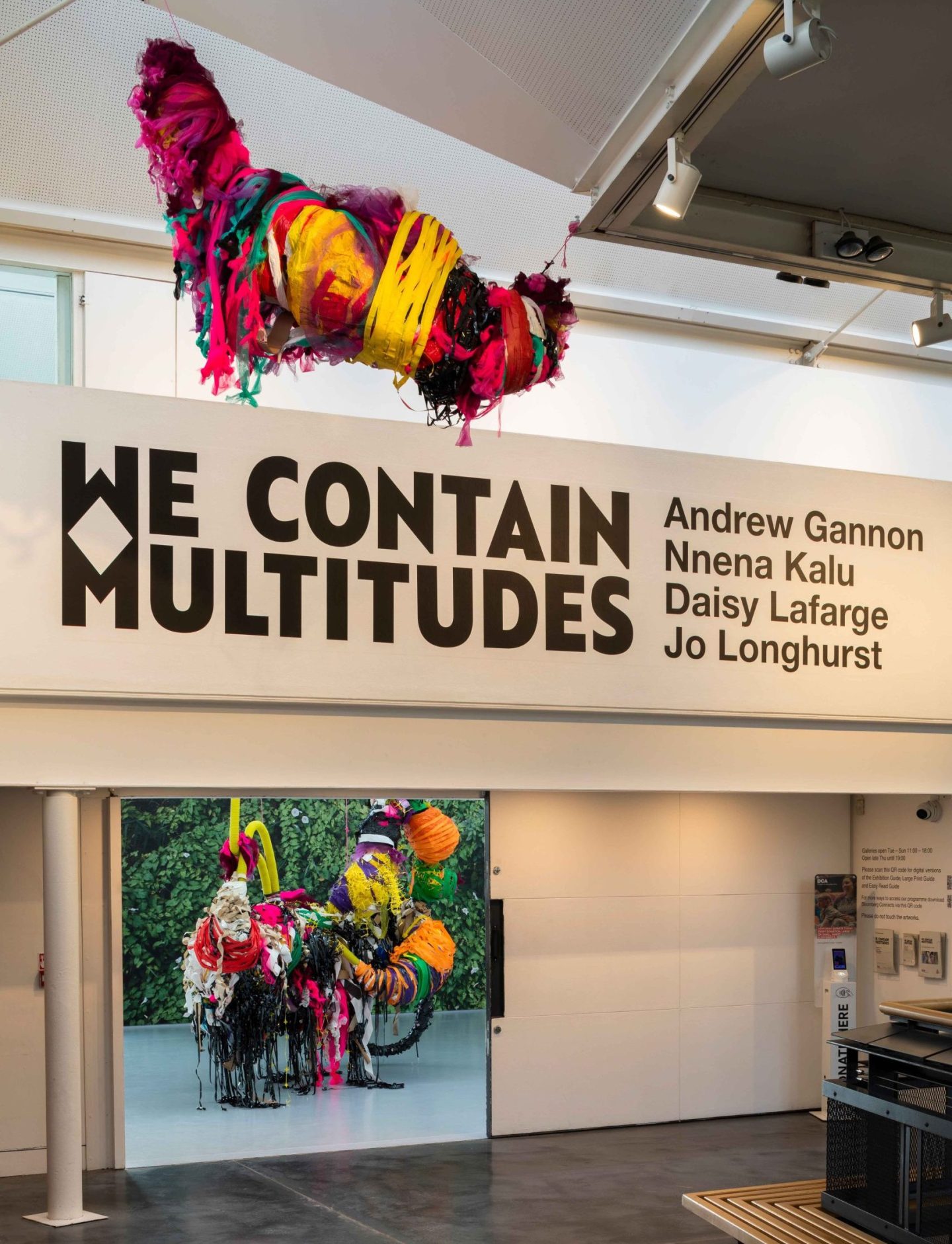 Colourful, large mixed media sculptures hung from a ceiling in a large exhibition space across two floors, large text on wall reads 'WE CONTAIN MULTITUDES'