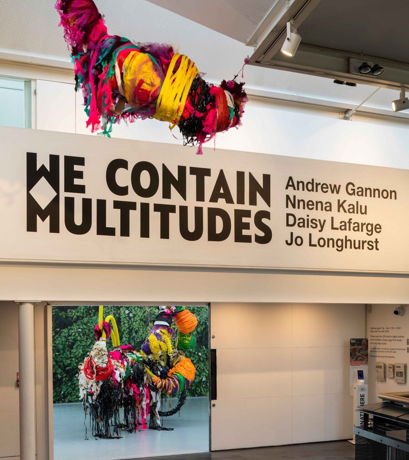 Colourful, large mixed media sculptures hung from a ceiling in a large exhibition space across two floors, large text on wall reads 'WE CONTAIN MULTITUDES'