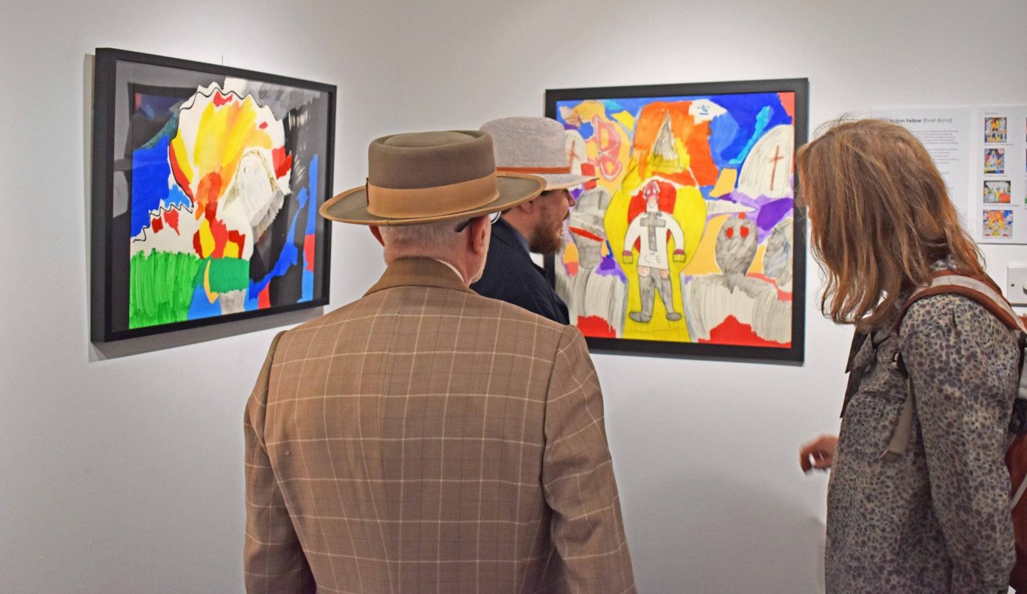 Image of three people looking at framed artworks hung on white walls. The works are graphic, coloured pencil drawings with political references.