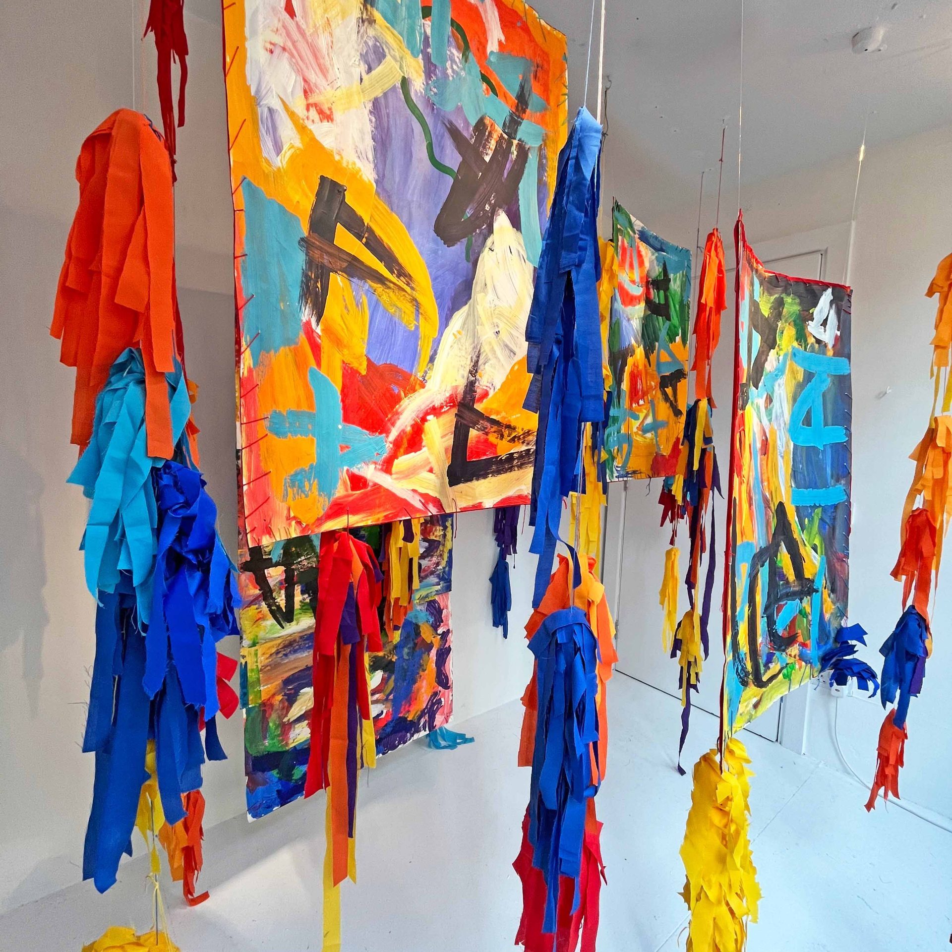 Image of an abstract installation with colourful textile ribbons and mixed media paintings hung from ceiling and installed in a small white gallery space.