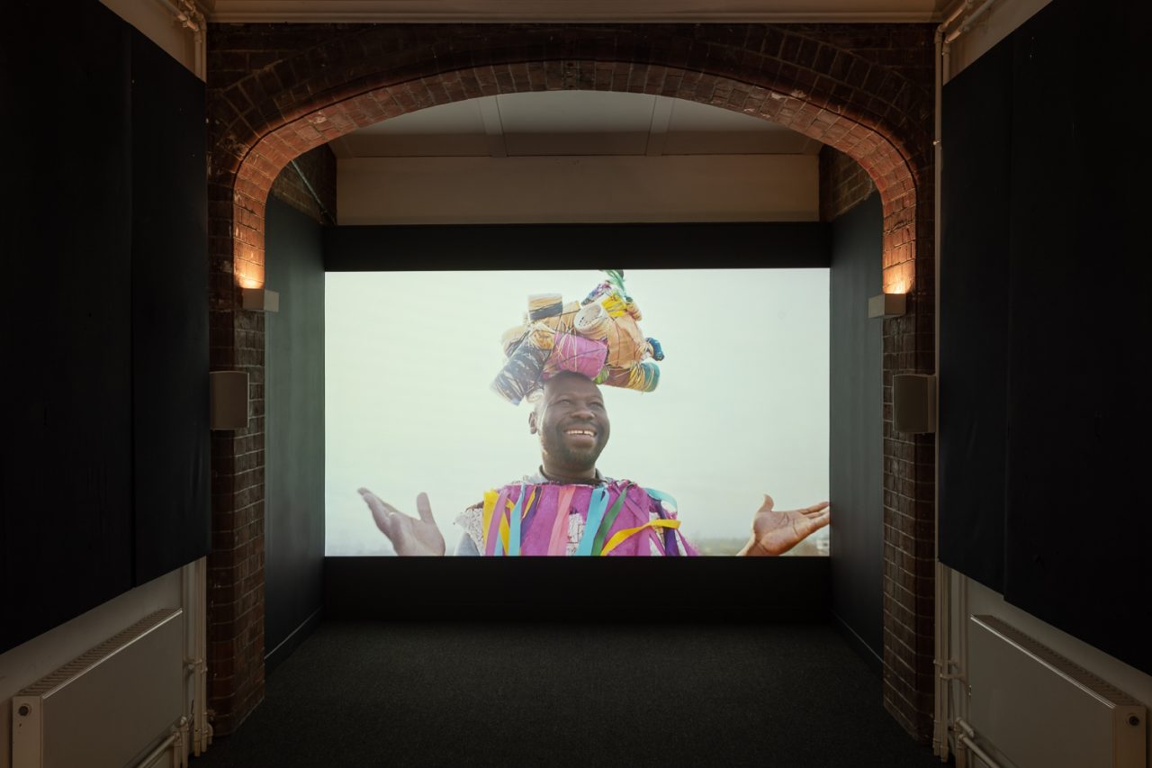 A still image from a film depicting a man smiling, wearing a colourful outfit.