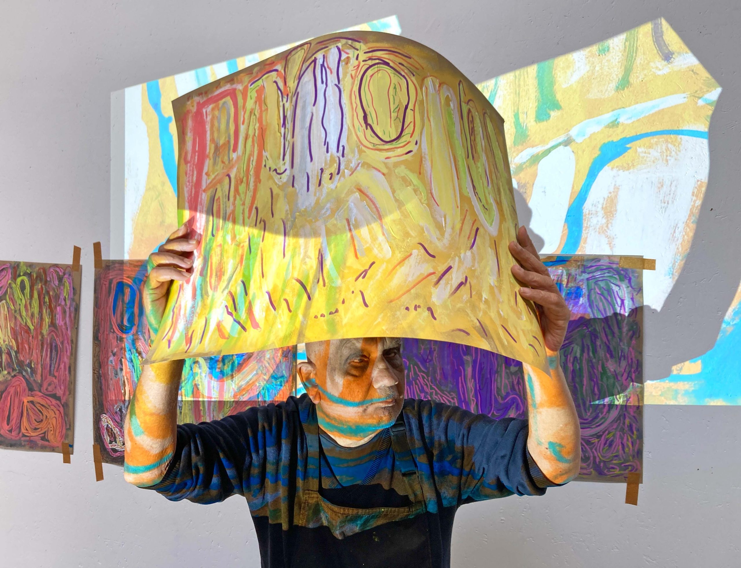 Artist Chandrakant Patel holding one of his paintings about his head. A projection of his artworks projects another artwork on top of himself, the artwork and the wall behind.