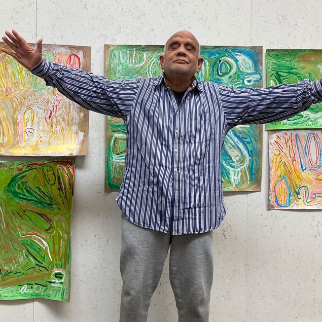 Artist Chandrakant Patel standing in front of a wall of his paintings. He is looking up with his arms raised up.