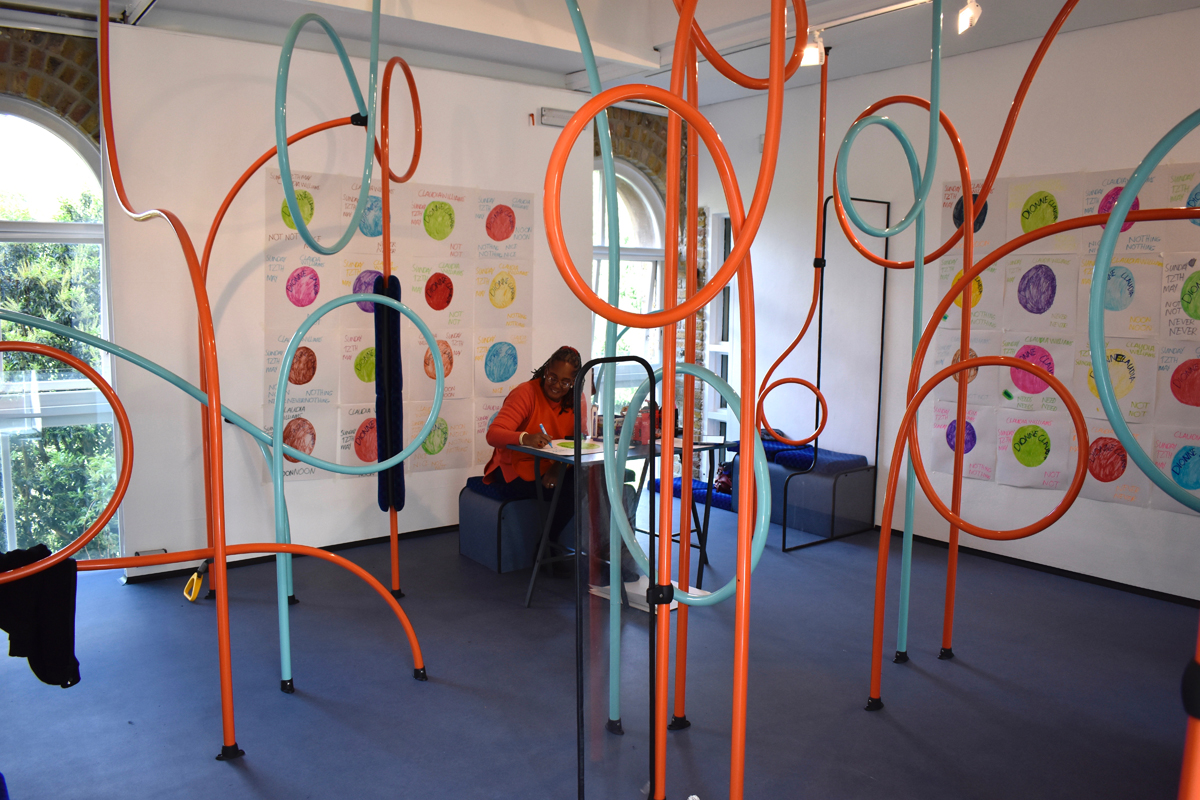 Artist Claudia Williams sat at a desk drawing within an exhibition installation at the Pump House Gallery.