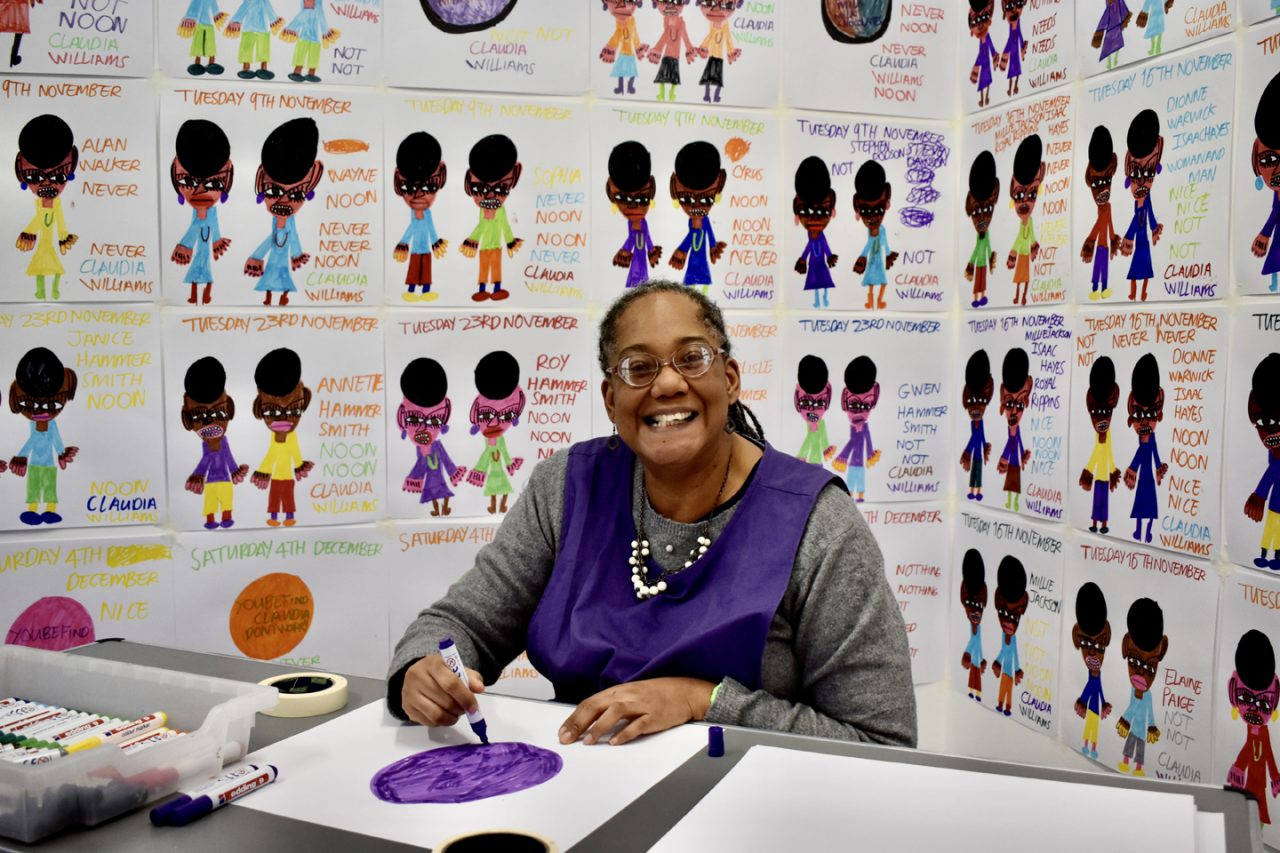 Artist Claudia Williams sat at a table drawing with her drawings installed behind her.