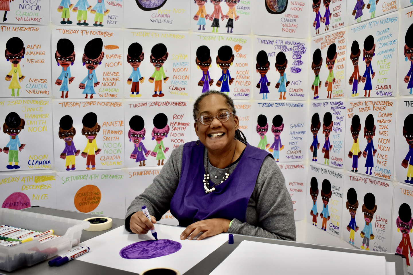 Artist Claudia Williams sat at a table drawing with her drawings installed behind her.