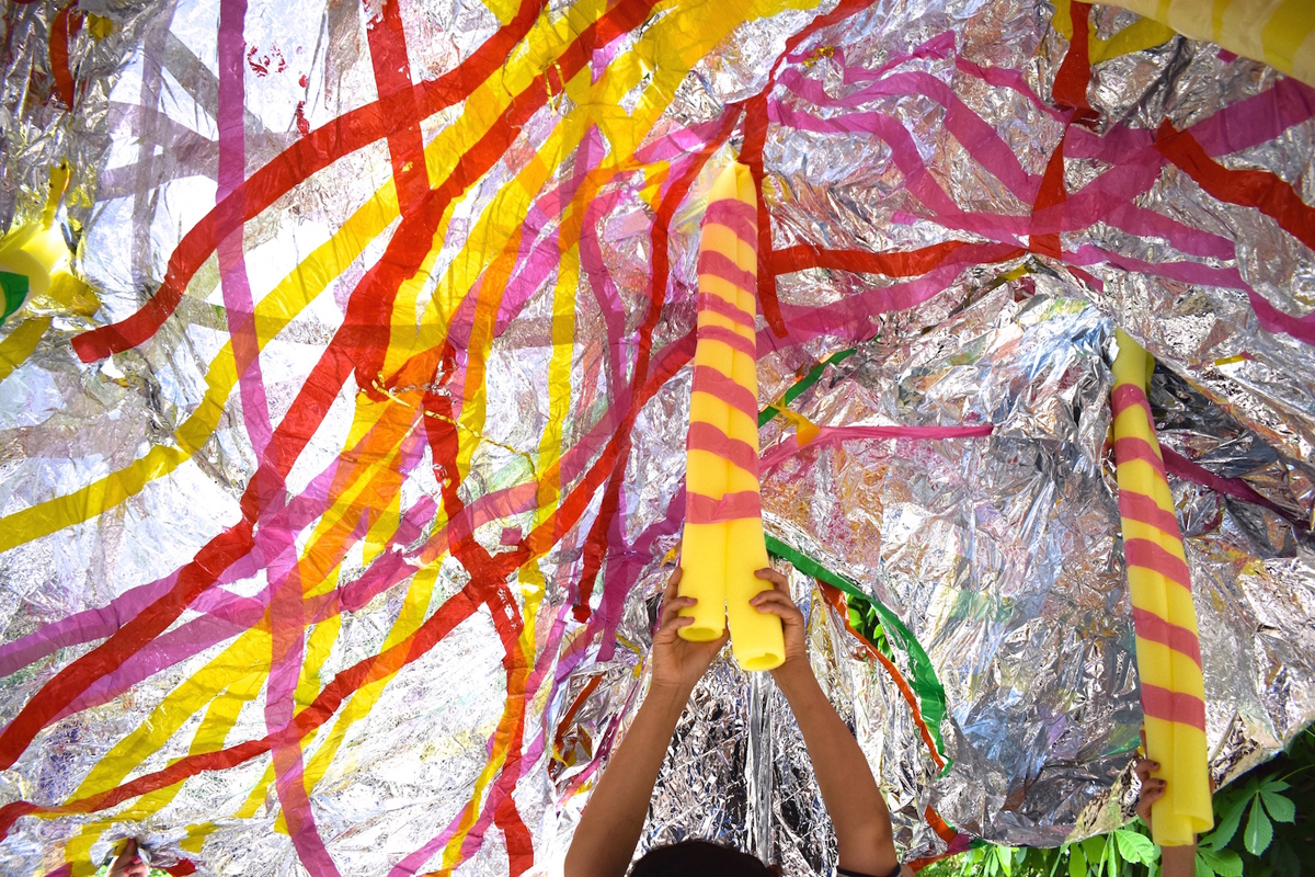 A silver foil sheet covered in red, yellow and pink tapes. Two yellow foam tubes are holding up the foil sheet, which two people are holding.