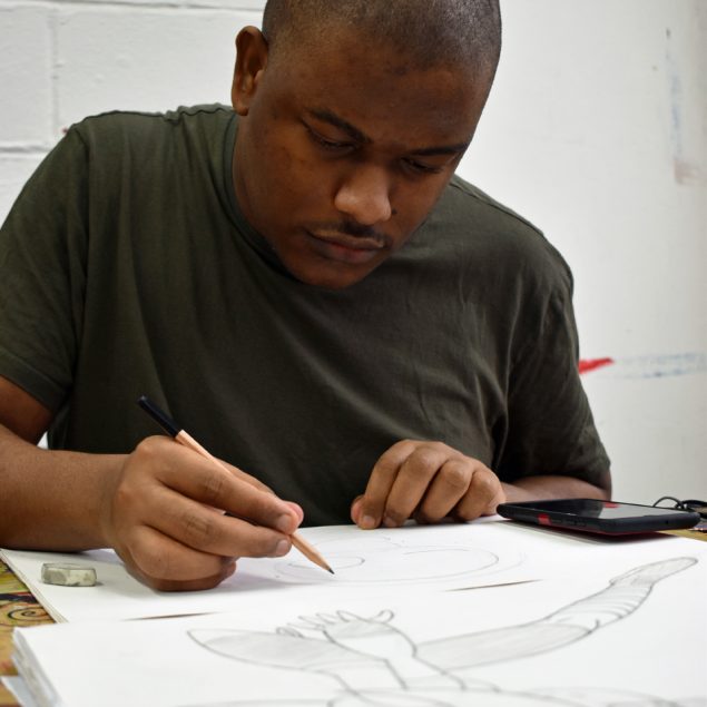 A man drawing in pencil on paper , sat at a desk.