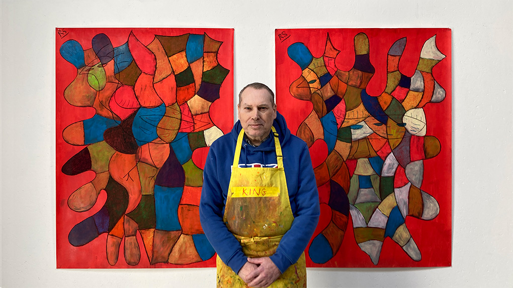 A male artist standing in front of two of his paintings that are abstract and red.