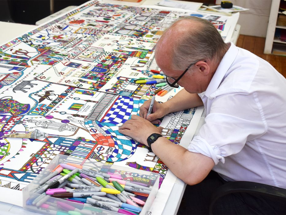 A man sitting at a table with a pen in his hand, working on a drawing.