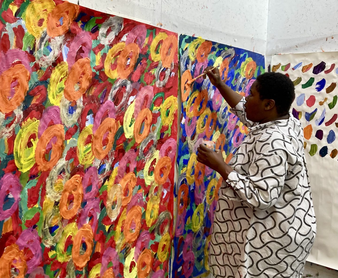 Artist Kwaga Sillingi standing up and painting on a large painting fixed to the wall.
