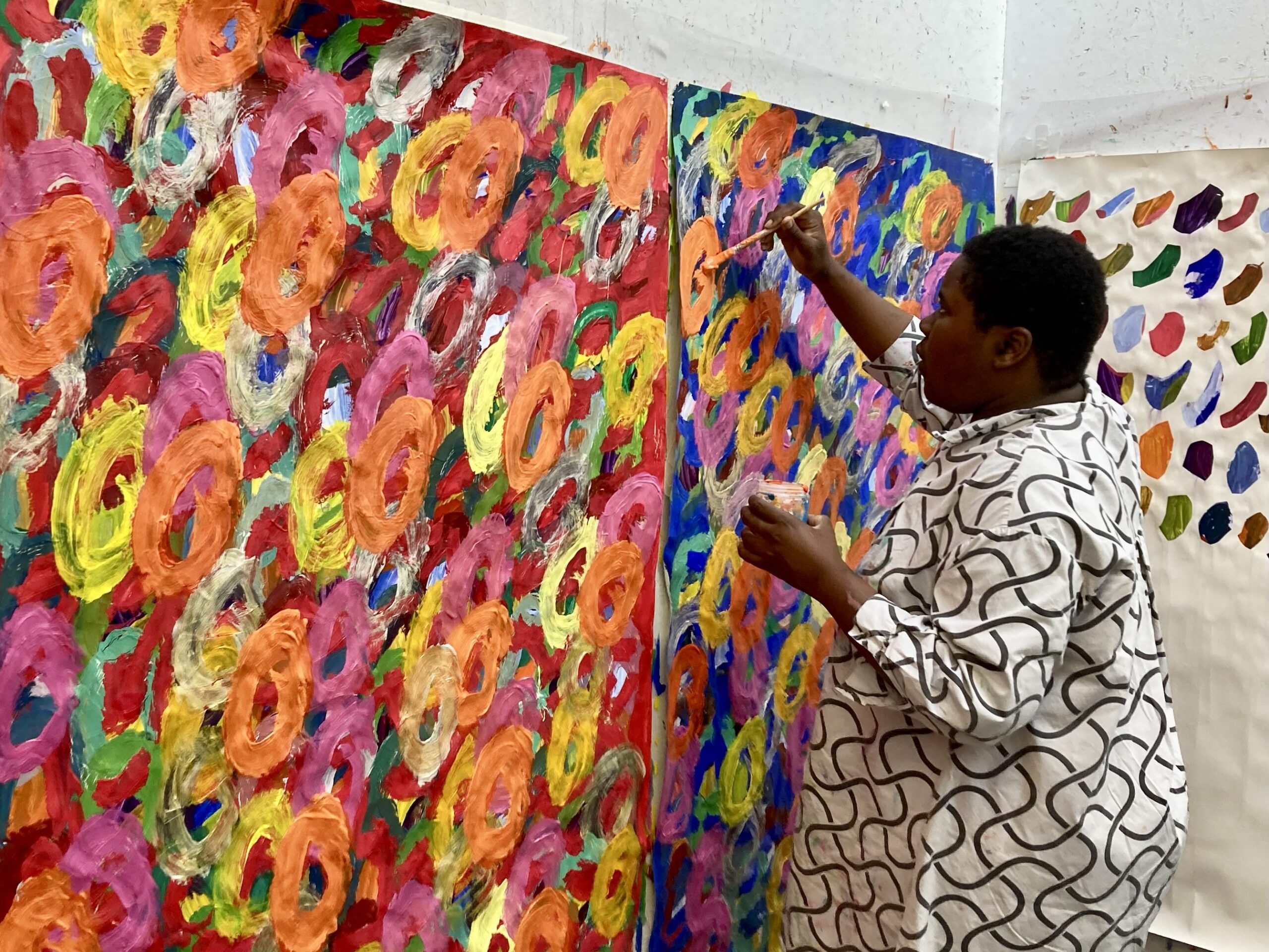 Artist Kwaga Sillingi standing up and painting on a large painting fixed to the wall.