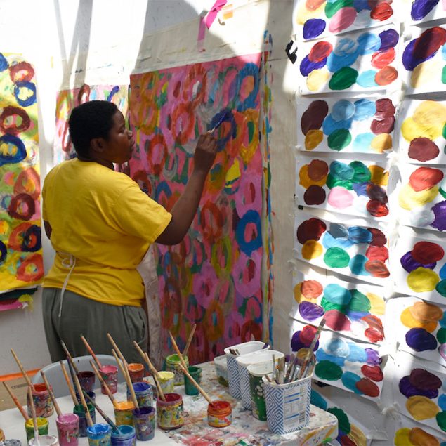 Artist Kwaga Sillingi standing up painting a painting that is attached to the studio wall.