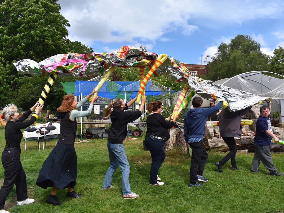Seven people walking in a line in a garden. They are holding up a sculpture made out of foam tube and a silver metallic sheet.