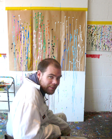 Declan is a white man with short dark hair and a short beard. He's wearing a white top and sitting in front of one of his paintings.