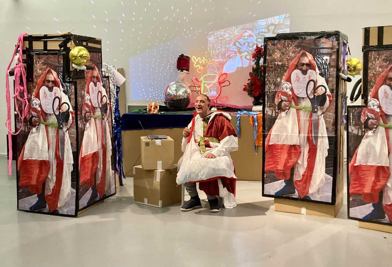 Pardip Kapul sat in the middle wearin a custom festive outfit. Either side of him are carboard boxes stacked on top of one another with large scale print outs of Pardip stuck onto them.