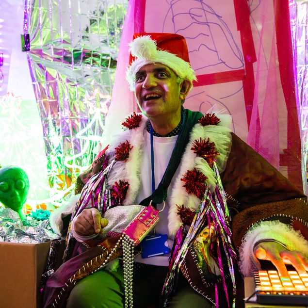 Pardip Kapil wearing a Father Christmas hat and custom made festive jacket. He is sat in his installtion at Studio Voltiare, surrounding by a silver metallic background, cardboard boxes and props.