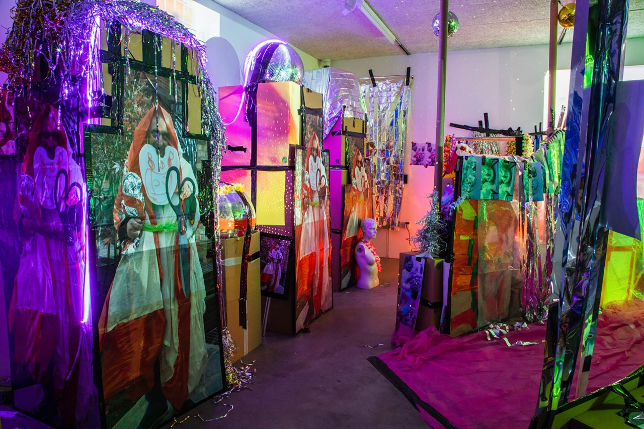 Installation of cardboax boxes with large photos of Pardip Kapil on, metallic strips of materials hanging and light projections on top of the installation.