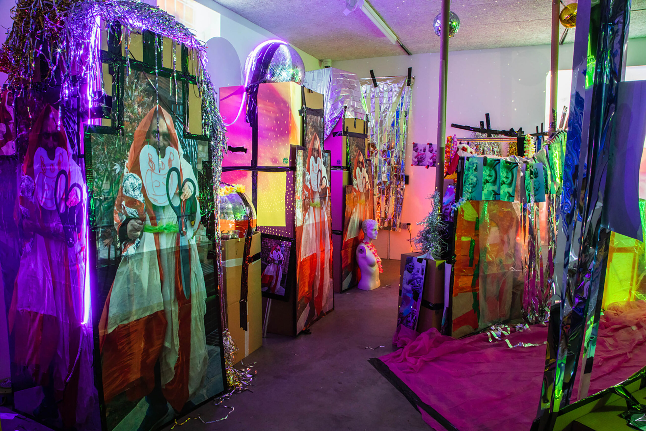 Installation of cardboax boxes with large photos of Pardip Kapil on, metallic strips of materials hanging and light projections on top of the installation.