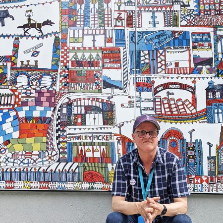 A picture of Ian Wornast. He is seated in front og his large colourful mural in Kingston. He is a white man and is wearing a blue har, glasses and a checked shirt and he has a blue lanyard around his neck.