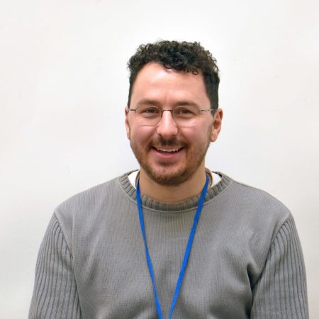 A picture of Louis Caseley. He is a white man with short curly hair. He has a closely cropped beard and he is wearing glasses, a grey jumper and has a blue lanyard around his neck.
