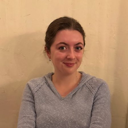 A picture of Maisie Rennie-Carter. She is a white woamn with medium length brown hair which has been tied behind herhead. She is wearing a grey jumper.