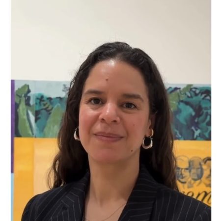 A photo of Dominique Hetse-Moore. She is w oman of mixed heritage with long dark hair. She is wearing a pint striped blazer and there are colourful paintings beind her.