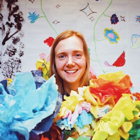 A photo of Jess Starns. She is a white woman with shoulder length, light brown hair. She is pictured emerging from a lot of colourful paper objects, there is a drawing on the wall behind her.