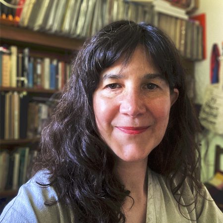A picture of KAte Squires. She is a white woman with long black hair. She is pictured on front of a book case and she is wearing a light coloured top.