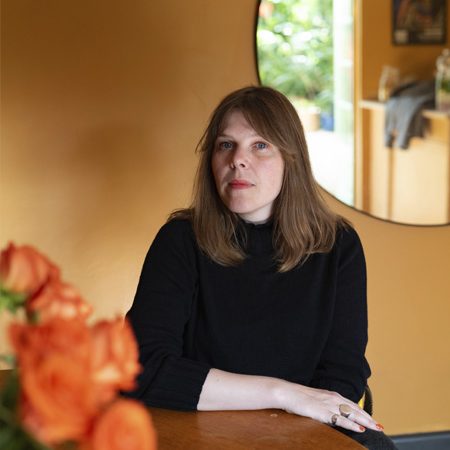 A photo of Linsey Young. She is a white woman with shoulder length brown hair. She isd wearing a dark jumper and is seated as table with a vase of flowers. There is a mirror on the wall behind her.