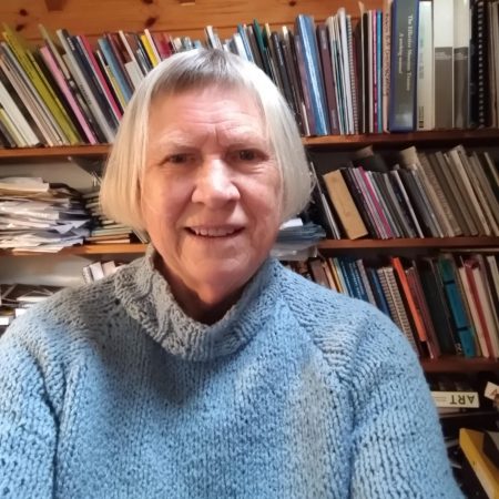 A picture of Loevday Shewell. She is a white woman with mid length grey hair. She is pcitrued in front of a book case and is wearing a blue jumper.