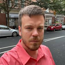 A picture of Sam Talbot. He is a white man with short brown hair, he has a closely cropped beard and is wearing a red shirt. He is standing in a street.