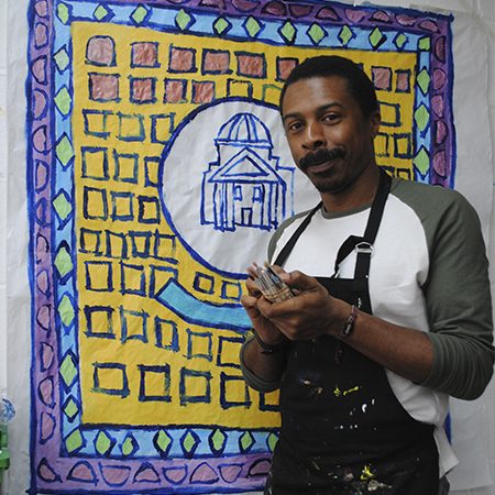 A picture of Thompson Hall standing in front of one of his paintings. He is a black man with a moustache and short hair and he is wearing a green and white t shirt and an apron.