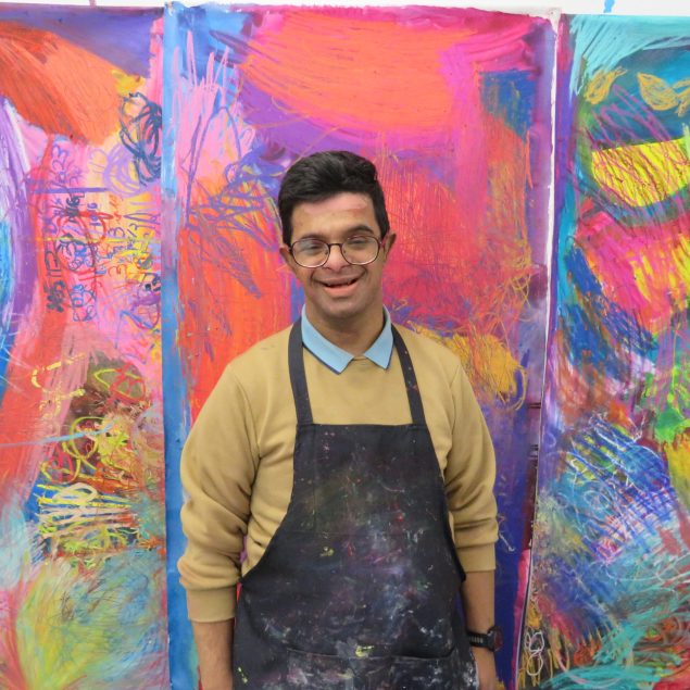 Artist Mohammed Imran Khan standing facing the camera smiling in front of his artwork. Hie is wearing a blue collared shirt, a beige jumper, and a navy artist's apron.