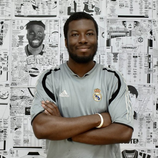 A picture of Babjide Brian. A black man with short black hair and beard. He is standing in front of a wall of his intrictate drawings of footballers and stadiums. He is wearing a football shirt.