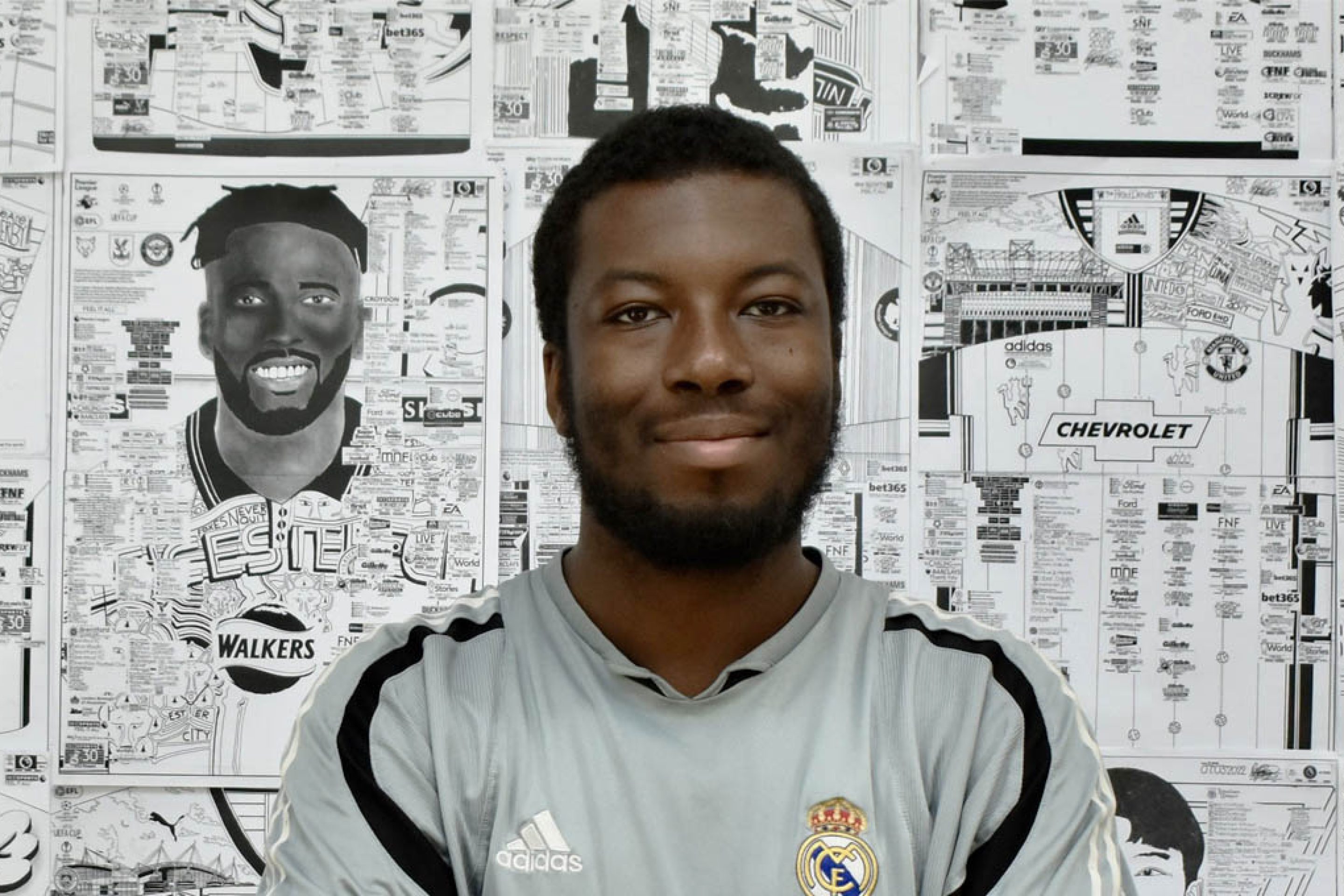 A picture of Babjide Brian. A black man with short black hair and beard. He is standing in front of a wall of his intrictate drawings of footballers and stadiums. He is wearing a football shirt.