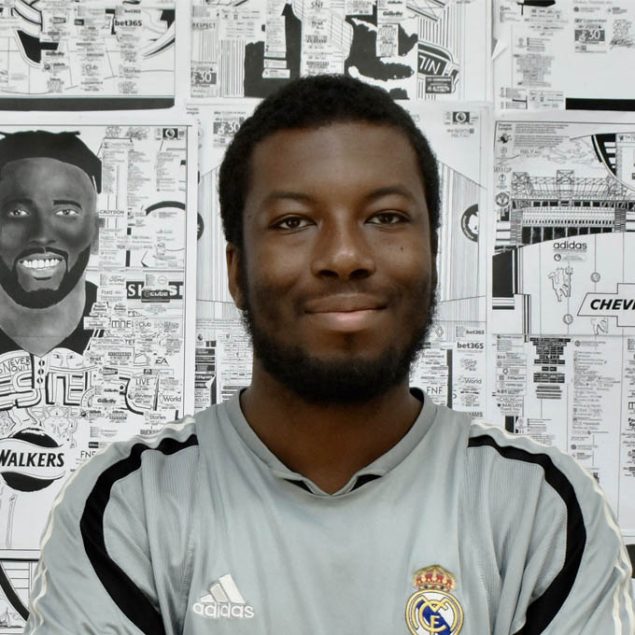 A picture of Babjide Brian. A black man with short black hair and beard. He is standing in front of a wall of his intrictate drawings of footballers and stadiums. He is wearing a football shirt.