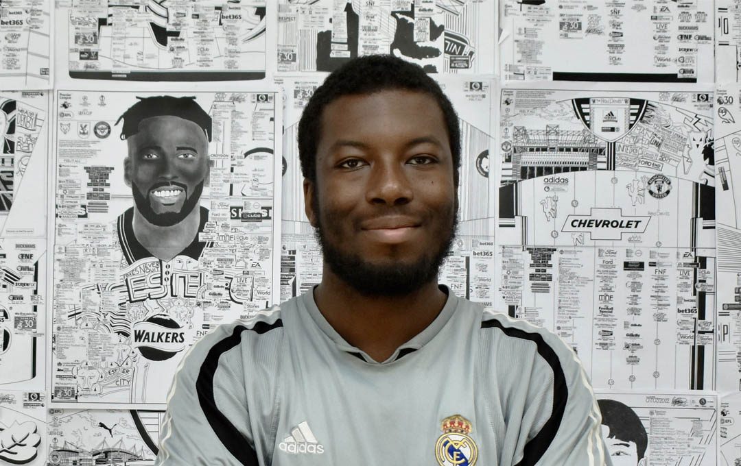 A picture of Babjide Brian. A black man with short black hair and beard. He is standing in front of a wall of his intrictate drawings of footballers and stadiums. He is wearing a football shirt.