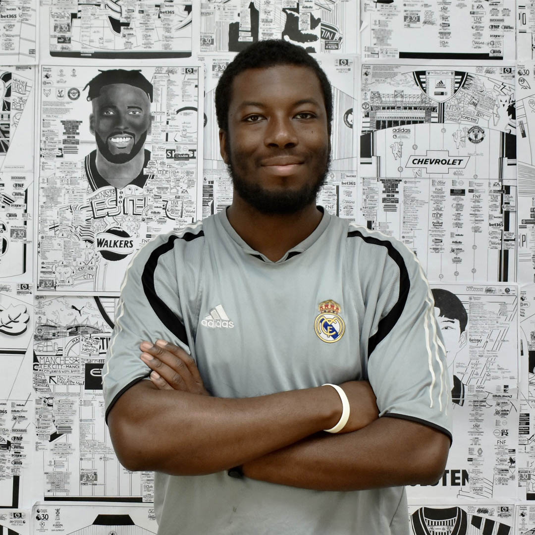 A picture of Babjide Brian. A black man with short black hair and beard. He is standing in front of a wall of his intrictate drawings of footballers and stadiums. He is wearing a football shirt.