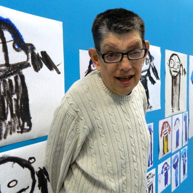 A picture of Roland Young. He is a white man with short dark hair and is wearing glasses and a cream knitted jumper. He is standing in front of several of his paintings which are displayed on a blue wall.