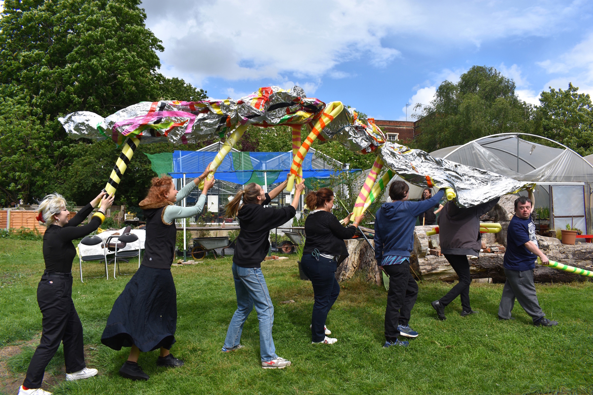 Seven people walking in a line holding up a silver foil sheet above their heads. They are holding onto yellow foam tubes that are propping up the foil sheet.