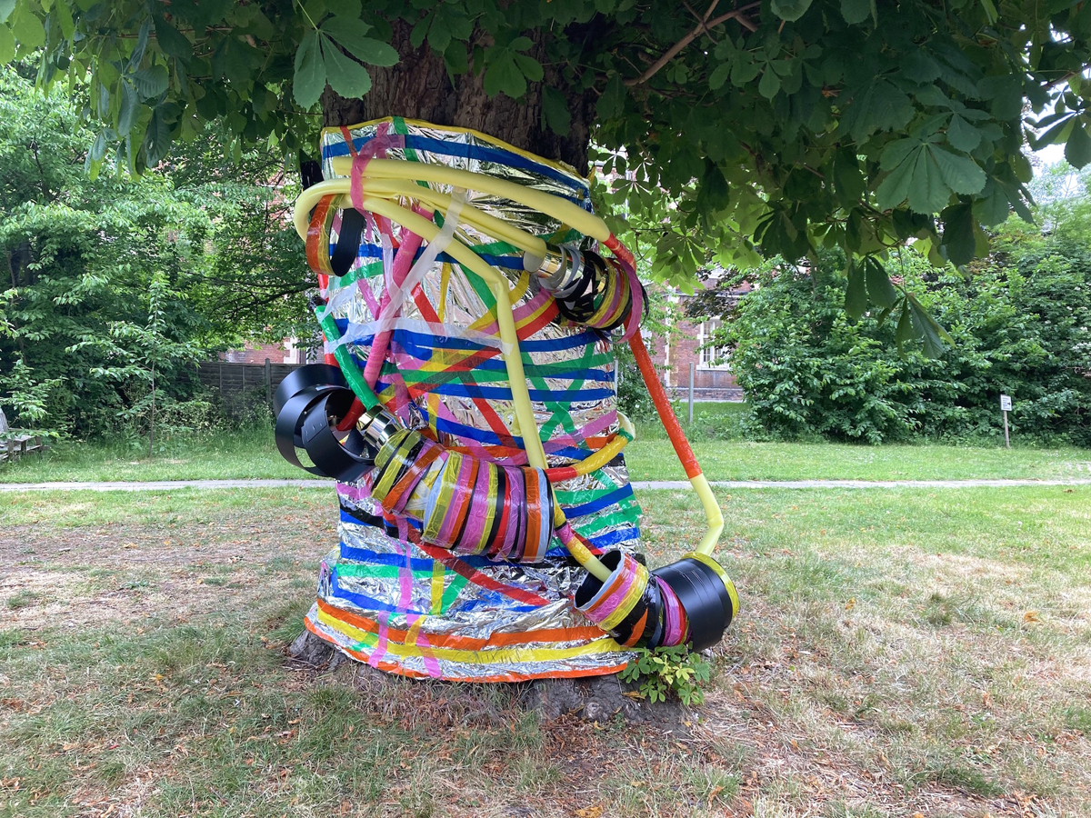 A large tree in the centre of the photo. Wrapped around the tree trunk is a foil sheet covered in coloured tape. Hanging from the tree are a series of long sculptures with hoop like parts hanging down.