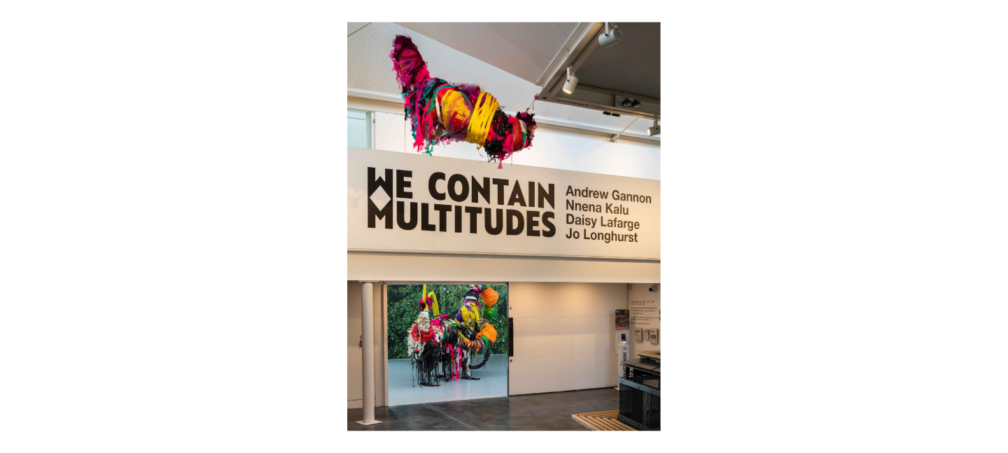 Colourful, large mixed media sculptures hung from a ceiling in a large exhibition space across two floors, large text on wall reads 'WE CONTAIN MULTITUDES'