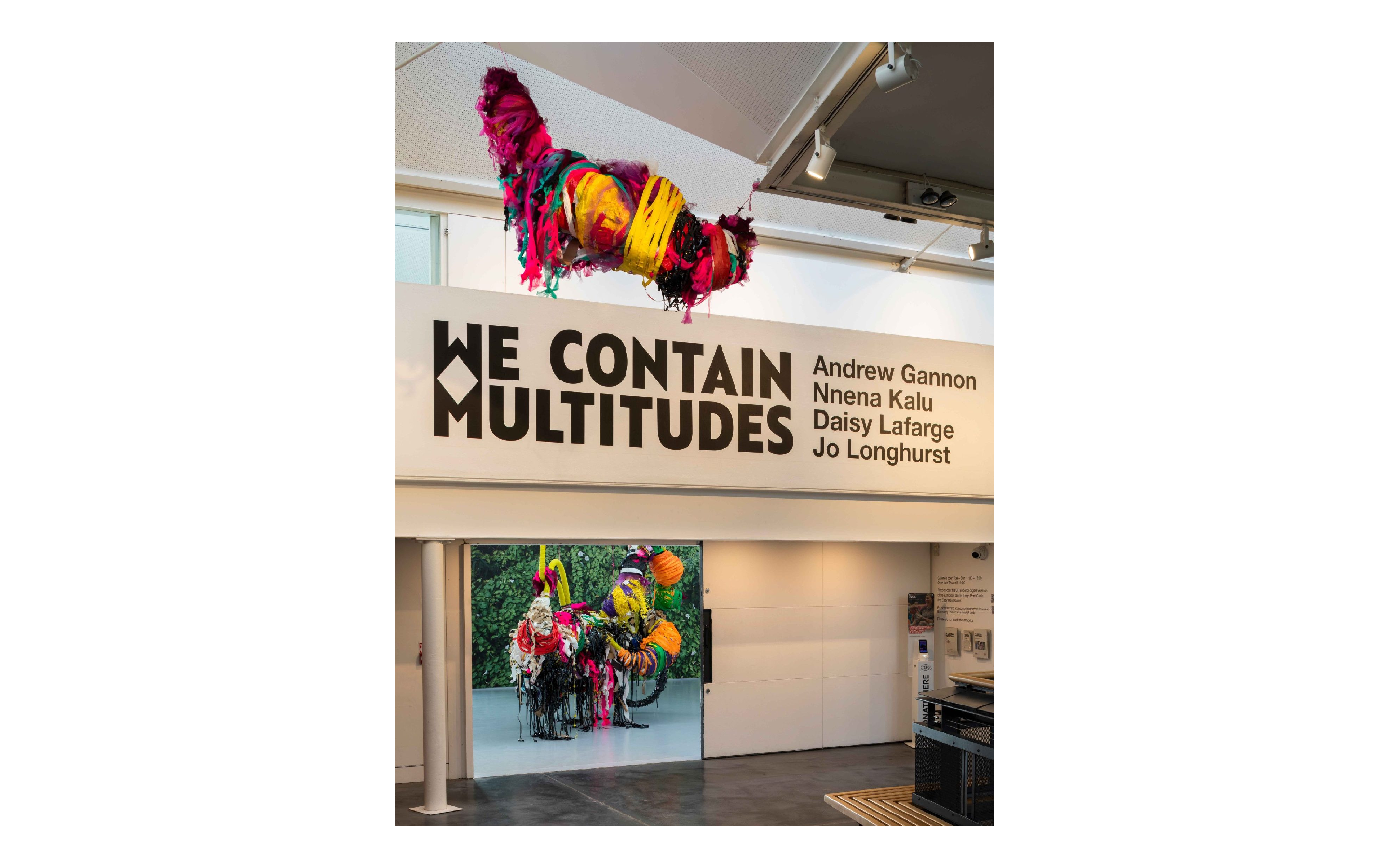 Colourful, large mixed media sculptures hung from a ceiling in a large exhibition space across two floors, large text on wall reads 'WE CONTAIN MULTITUDES'