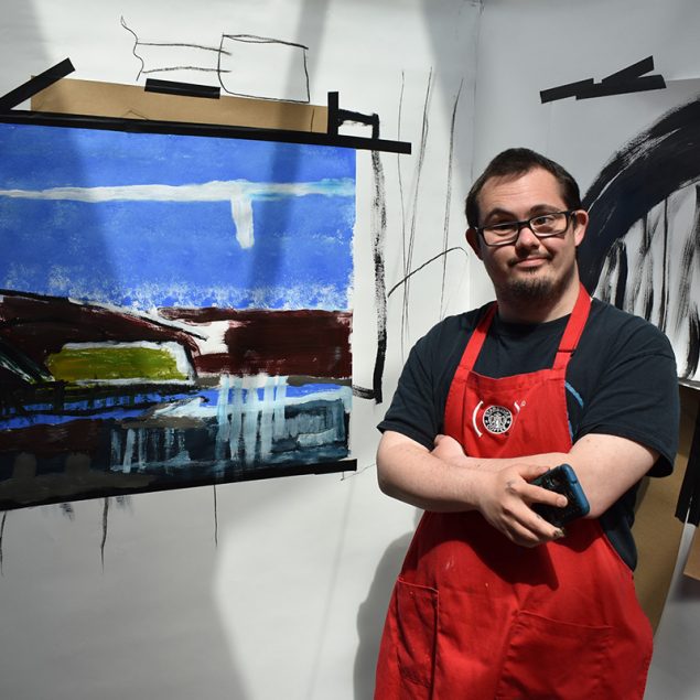 The artist James Heath stands next to two of his paintings, he is wearing glasses and a red apron and is holding a phone.