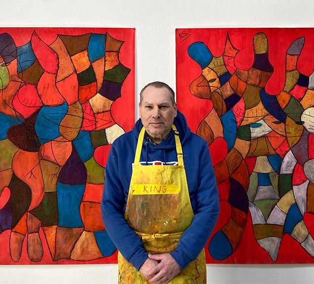 Artist Robin Smith standing in from of two paintings. He is wearing a yellow apron.